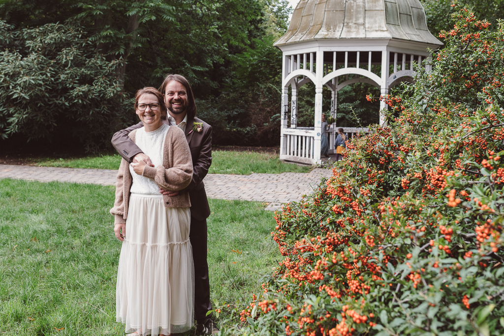 Brautpaar im Johannapark beim Portraitshooting. Der Mann umarmt seine Frau von hinten, im Hintergrund sieht man einen weißen Pavillon.