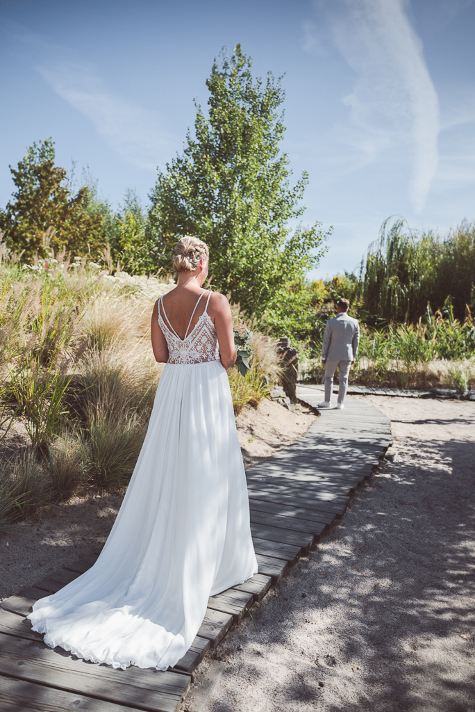 First Look. Braut im Vordergrund in Rückenansicht, im Hintergrund sieht man den Bräutigam, ebenfalls in Rückenansicht. Beide stehen auf einem Holzsteg im Sand.