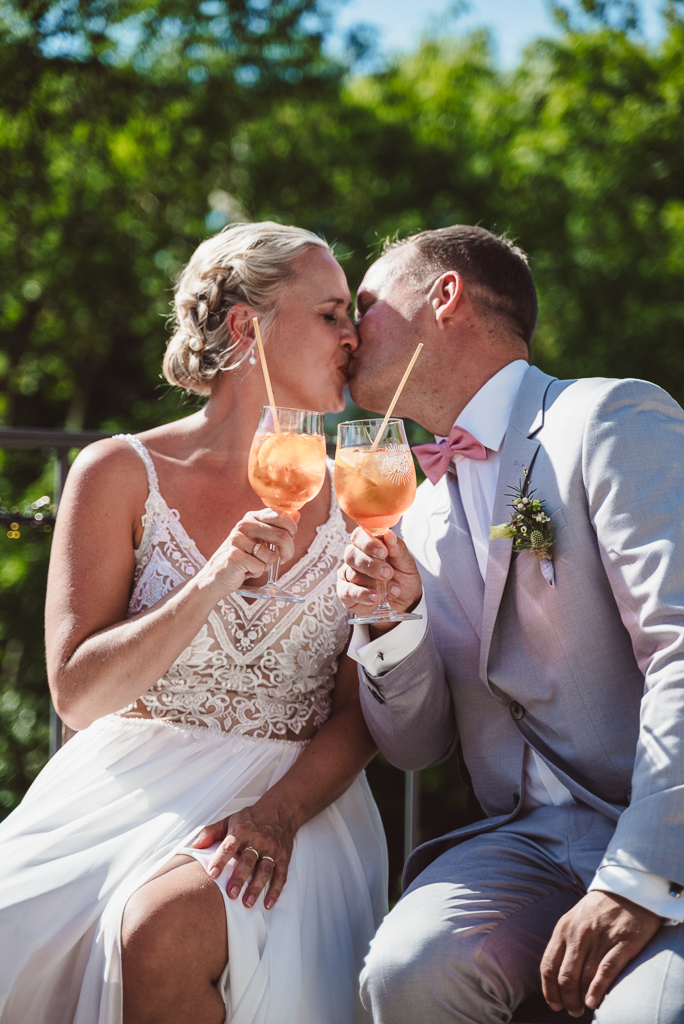 Sitzendes Brautpaar gibt sich einen Kuss. In ihren halten sie jeweils ein Glas Aperol Sprizz.