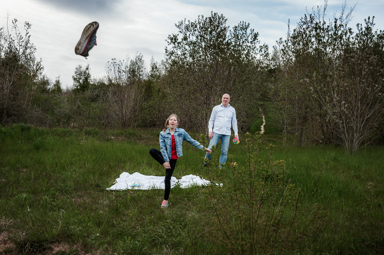 Familie spielt auf einer Decke im Gras für ungestellte Familienfotos in der Natur und ein Schuh fliegt durch die Luft.