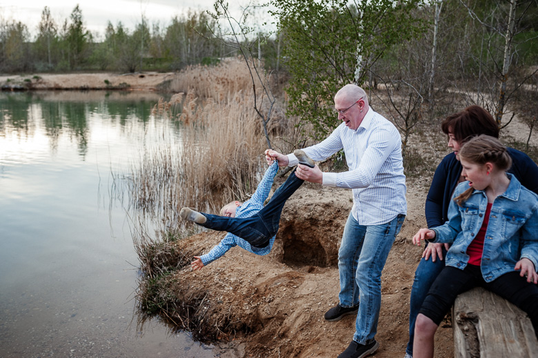 Familie albert am See, der Vater hält den Sohn an Arm und Bein und wirbelt ihn über das Wasser für ungestellte Familienfotos am see.